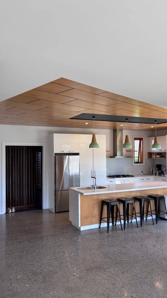 Modern kitchen with a long island, sink, and black bar stools under a wood-paneled ceiling. pendant lights glow over white cabinets and stainless fridge.