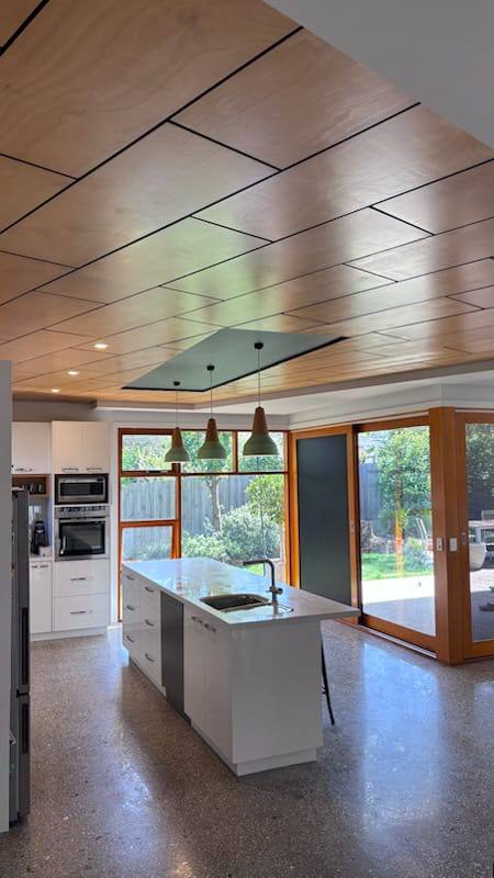 Modern kitchen with white island, built-in ovens, and wood-paneled ceiling; three pendant lights hang above the island and glass doors open to greenery outside.